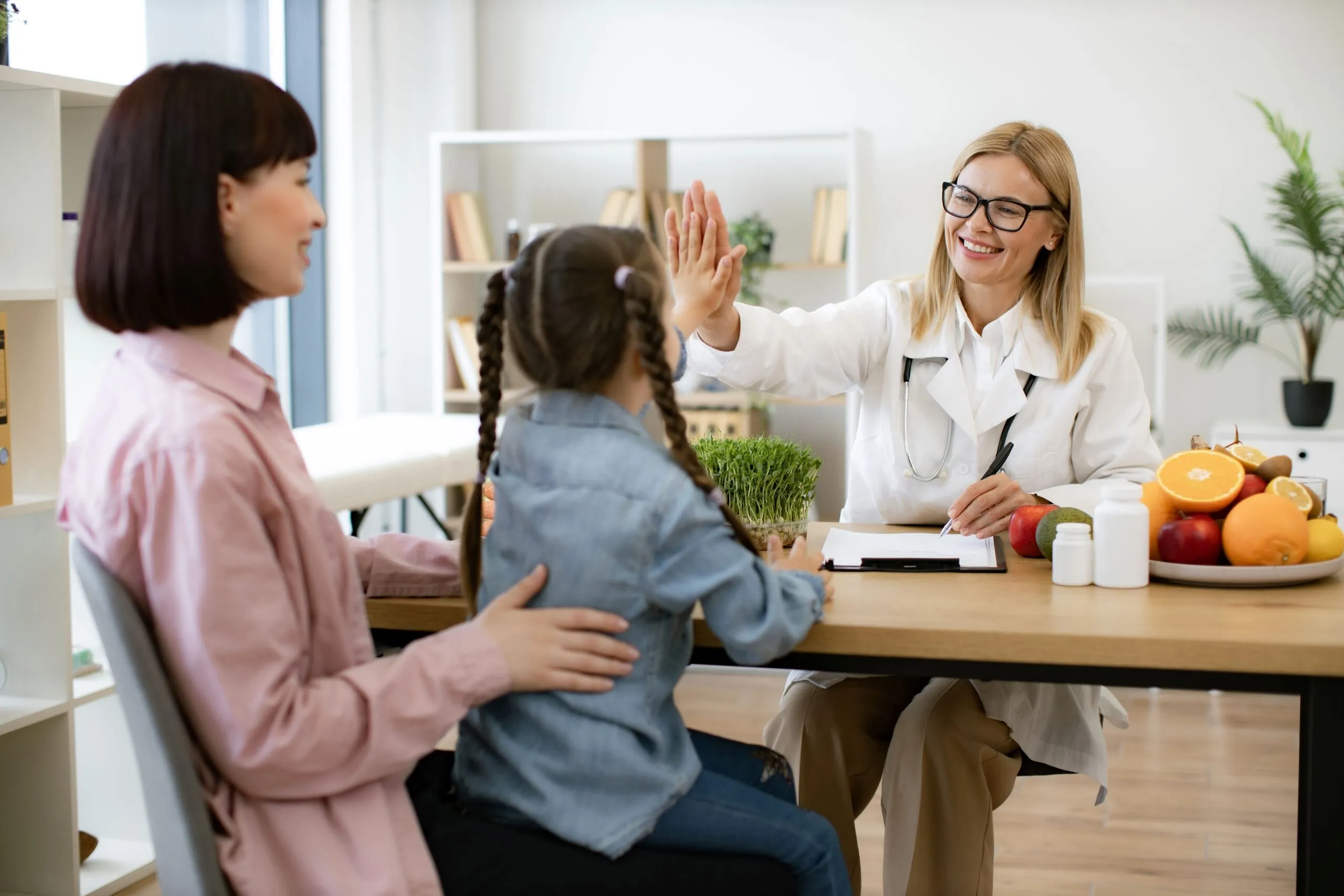 Doctora sonriente da un saludo de manos a una niña durante una consulta nutricional, con frutas frescas y suplementos sobre la mesa, mientras la madre acompaña a su hija.