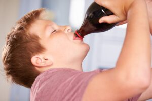 Niño bebiendo directamente de una botella de refresco oscuro, inclinado hacia atrás y con los ojos cerrados, en un entorno interior iluminado.