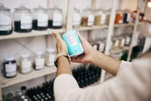 Primer plano de una persona sosteniendo un envase y leyendo su tabla nutricional en una tienda con estantes llenos de frascos de alimentos.
