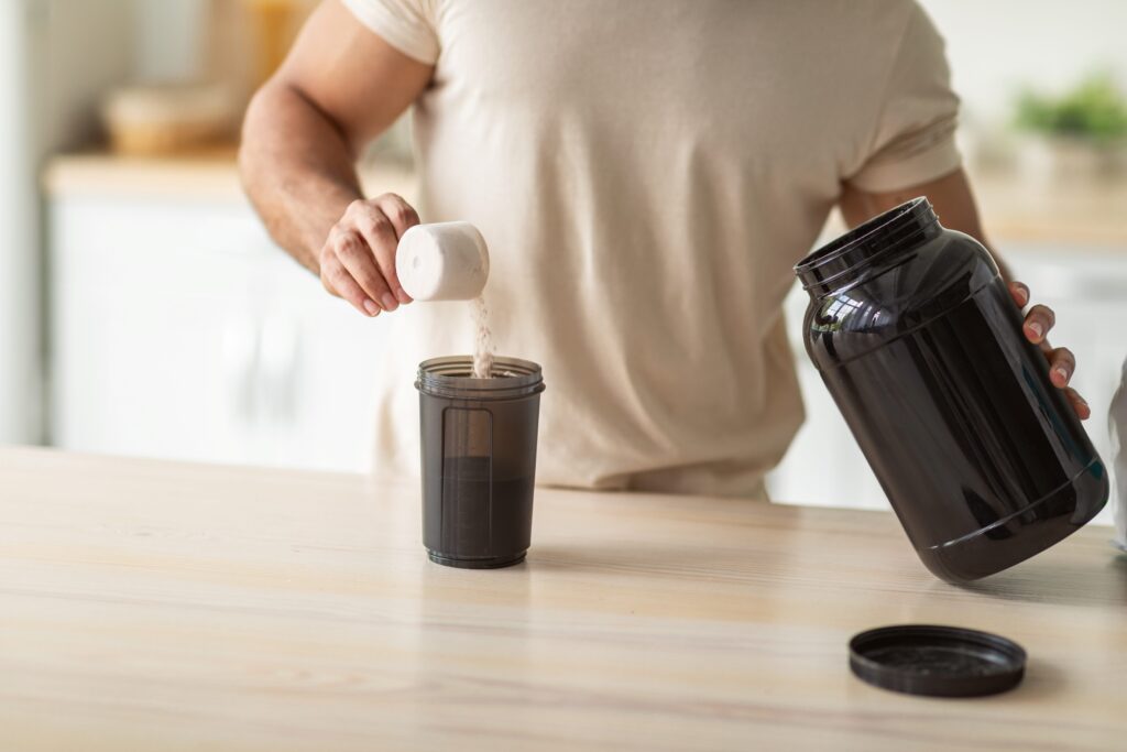 Hombre sirviendo proteína en polvo desde un bote negro a un vaso medidor, sobre una mesa de madera clara, en una cocina iluminada.