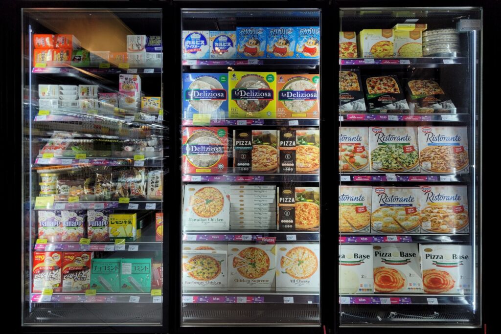 Vitrina refrigerada de supermercado con puertas de cristal, mostrando pizzas congeladas de distintas marcas, junto con otros productos congelados empaquetados, organizados en varias repisas iluminadas.