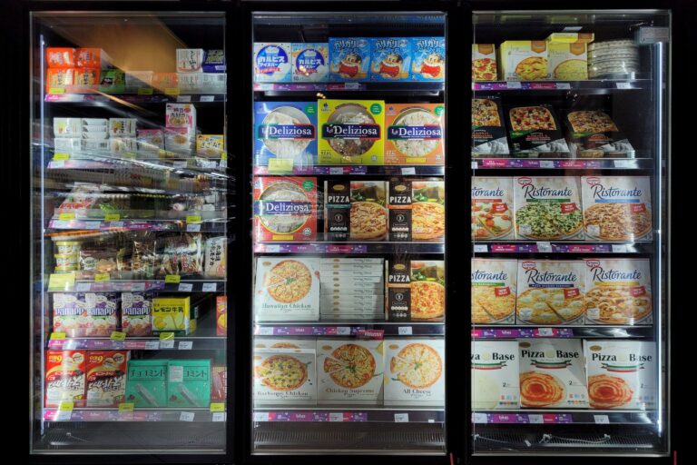 Vitrina refrigerada de supermercado con puertas de cristal, mostrando pizzas congeladas de distintas marcas, junto con otros productos congelados empaquetados, organizados en varias repisas iluminadas.