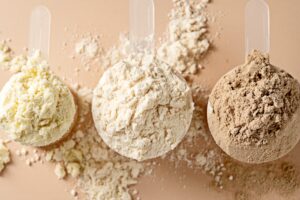 Tres cucharas medidoras con proteína en polvo de diferentes tonalidades —vainilla, natural y chocolate— sobre una superficie beige.