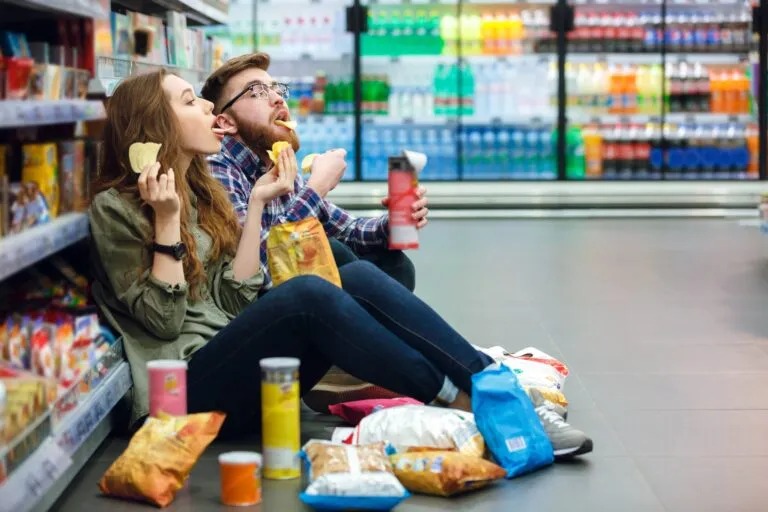 Pareja de jóvenes sentados en un pasillo de supermercado comiendo papas fritas y rodeados de botanas empaquetadas, representando el consumo de alimentos ultraprocesados y sus riesgos para la salud.