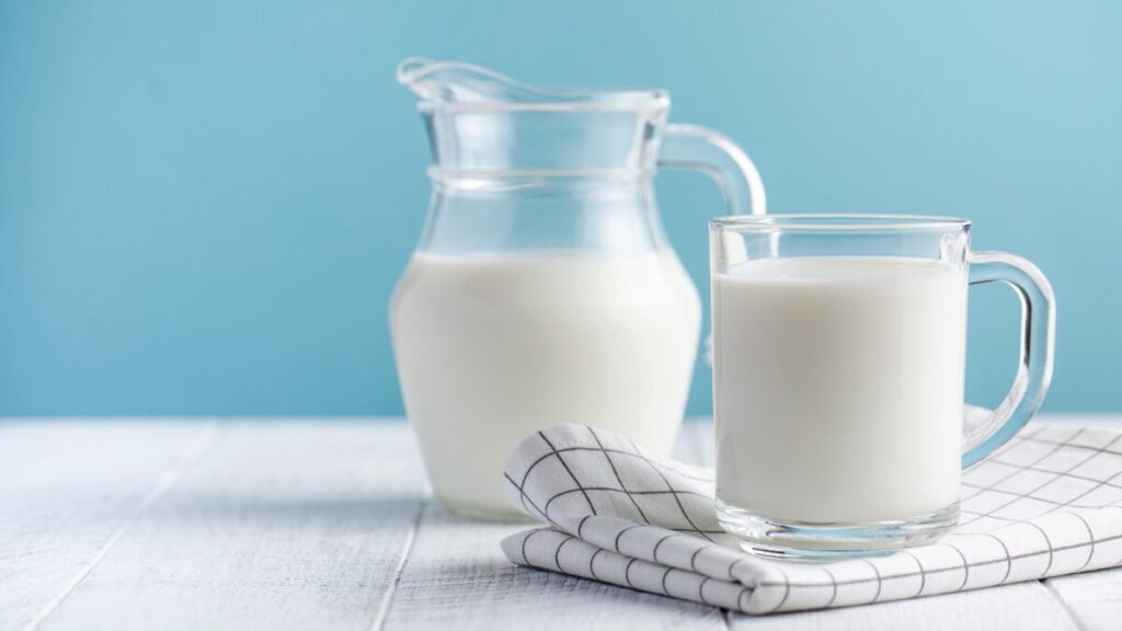 Vaso y jarra de leche fresca sobre una mesa blanca con fondo azul, representando el análisis de leche reprobada por Profeco.