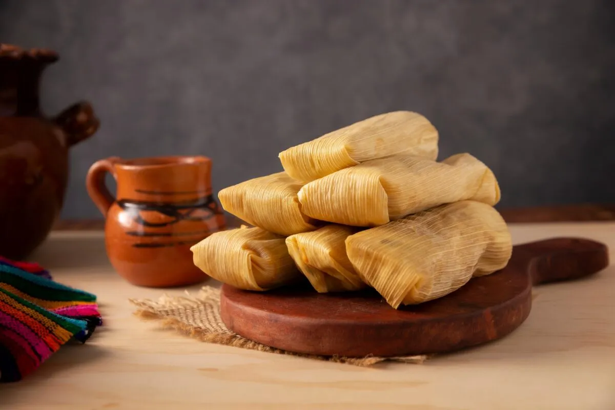 Tamales mexicanos envueltos en hoja de maíz sobre una tabla de madera, acompañados de una jarra artesanal y colores típicos mexicanos