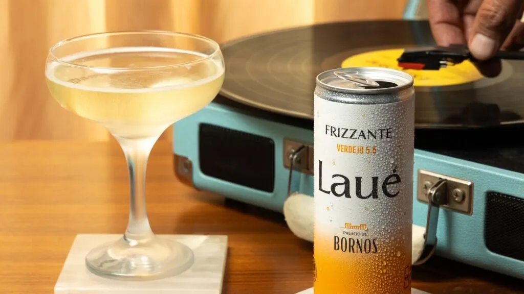 Lata de LAUÉ frizzante bien fría junto a una copa servida, colocadas sobre una mesa de madera. Al fondo, un tocadiscos reproduce un vinilo, creando una escena lifestyle relajada que evoca un momento de disfrute y celebración cotidiana.