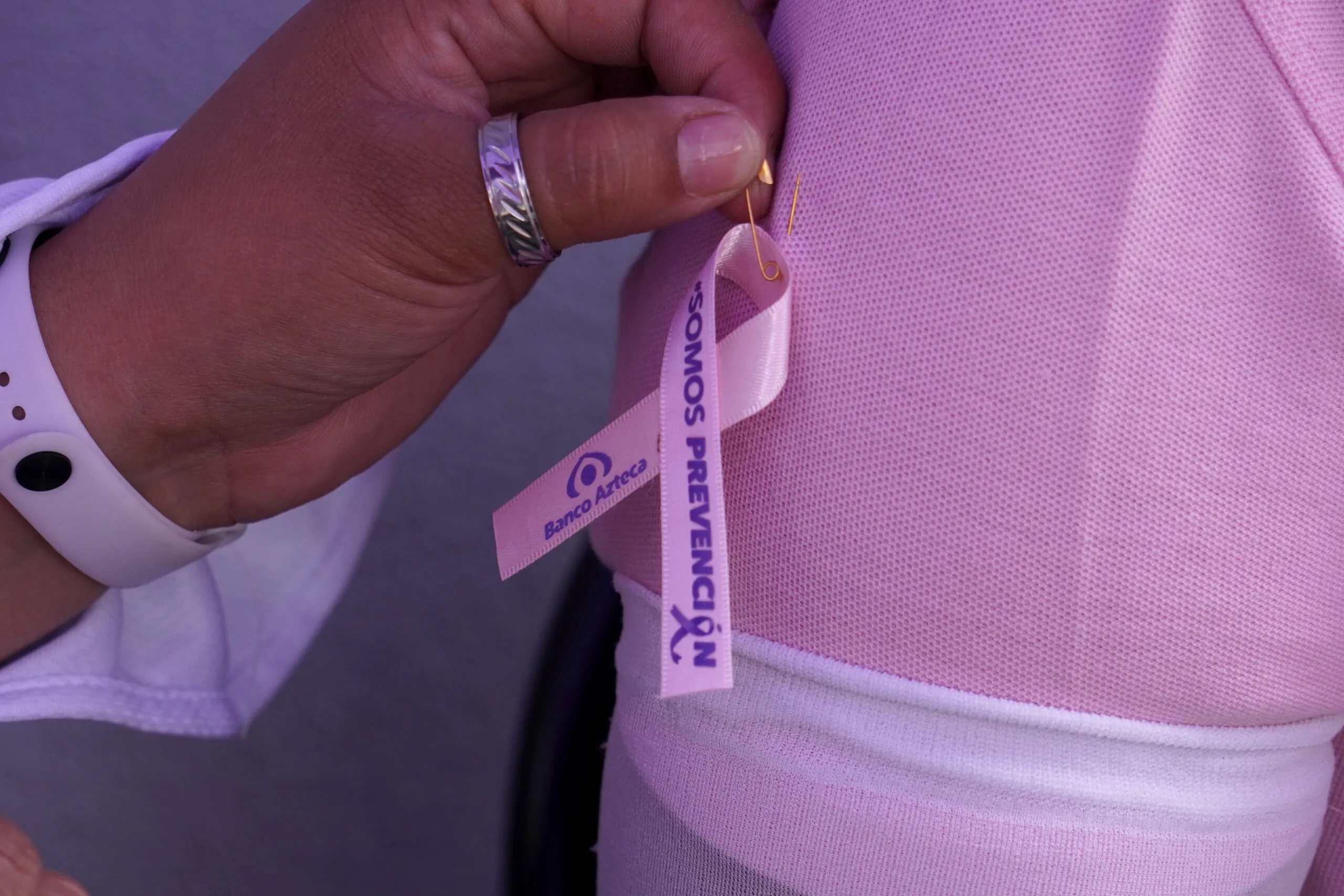 Mano coloca un listón rosa de la campaña SOMOS Prevención de Banco Azteca sobre una prenda rosa.