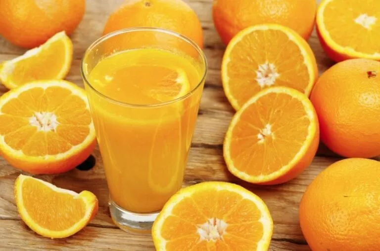 Vaso de jugo de naranja acompañado de naranjas enteras y partidas sobre una mesa